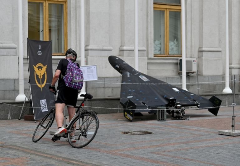 A man on a bicycle observes a downed black drone outside an aged structure